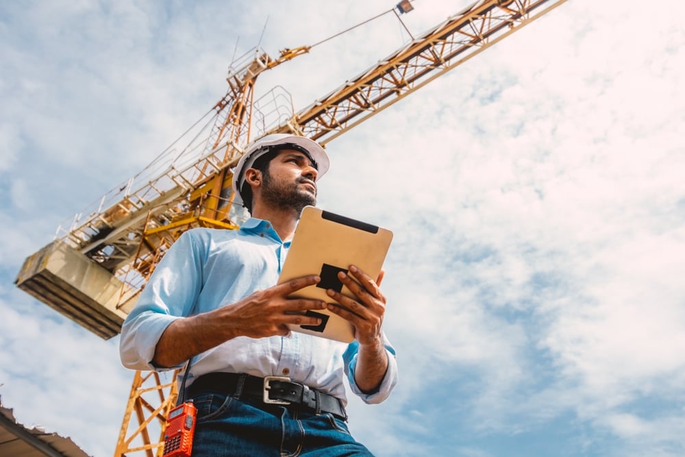 Construction worker in hard hat holds a clipboard while standing under a crane against a blue sky, surveying the site.